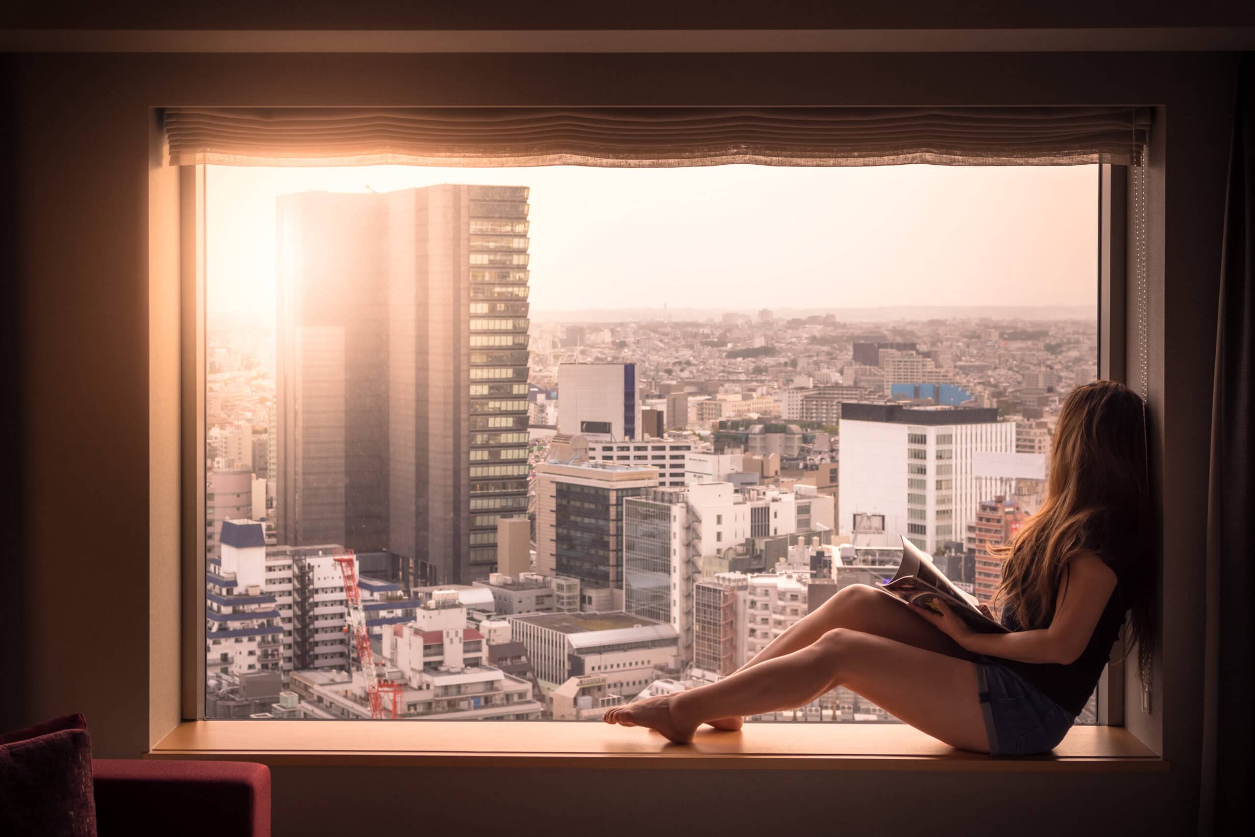 A woman is relaxed and looking out of her hotel window. She's looking at the cityscape of Shibuya, Tokyo, Japan below. Shot in the morning in Spring.